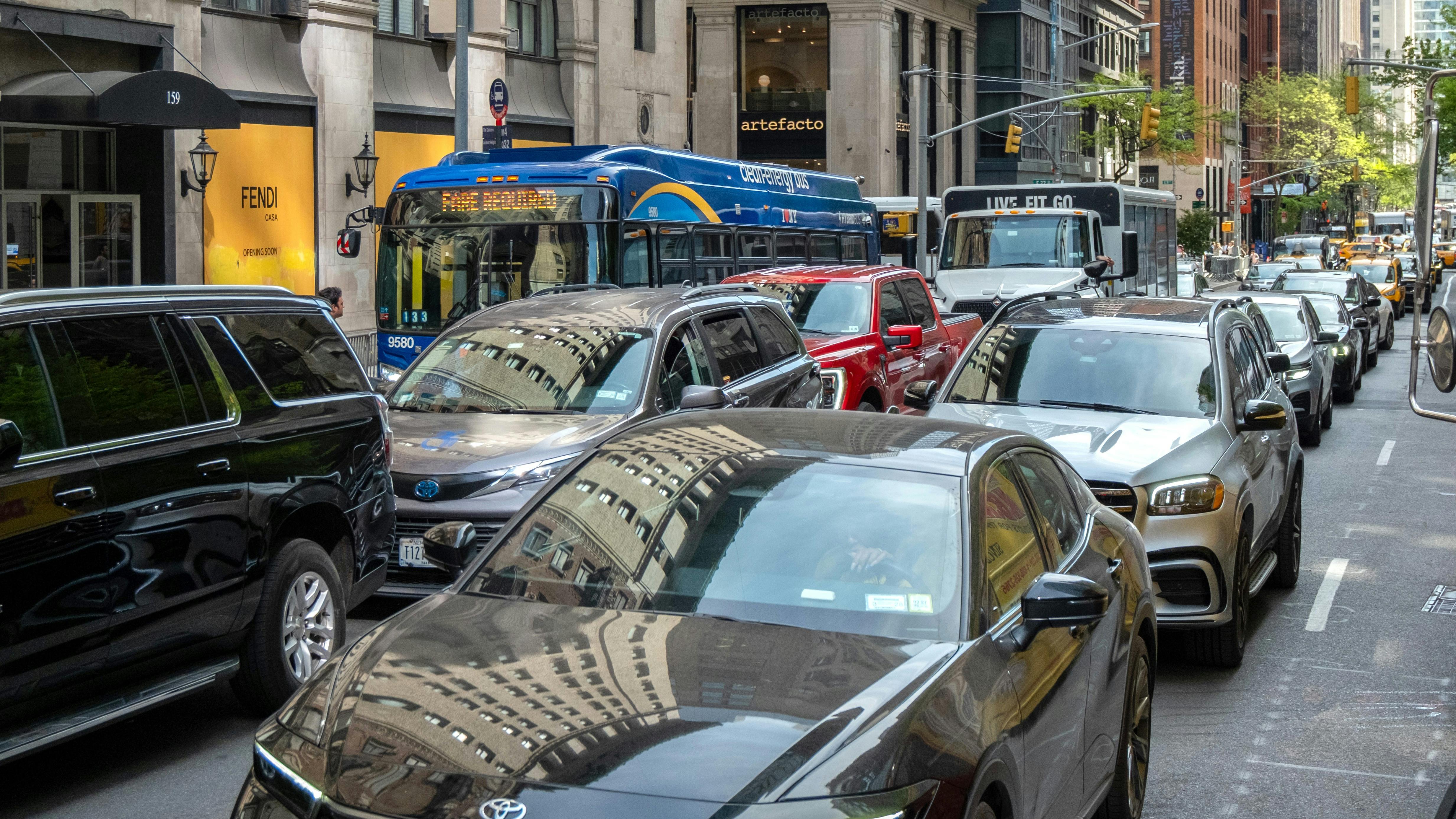 A New York City Transit bus and several cars sit in gridlocked traffic on Madison Avenue in Midtown Manhattan, with luxury storefronts including Fendi visible along the sidewalk.