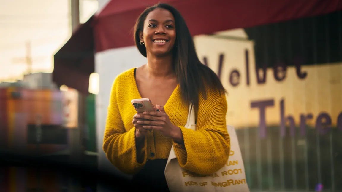 A smiling woman holding a smartphone and a canvas tote bag waits outside a storefront at what appears to be dusk, suggesting she may be waiting for a ride.