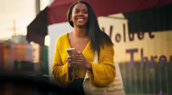 A smiling woman holding a smartphone and a canvas tote bag waits outside a storefront at what appears to be dusk, suggesting she may be waiting for a ride. A smiling woman holding a smartphone and a canvas tote bag waits outside a storefront at what appears to be dusk, suggesting she may be waiting for a ride.