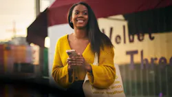 A smiling woman holding a smartphone and a canvas tote bag waits outside a storefront at what appears to be dusk, suggesting she may be waiting for a ride. A smiling woman holding a smartphone and a canvas tote bag waits outside a storefront at what appears to be dusk, suggesting she may be waiting for a ride.