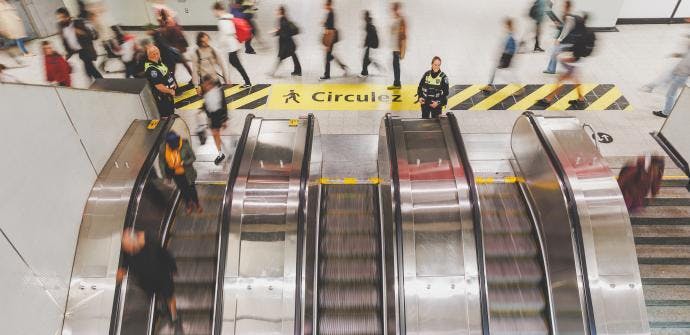 Two STM security officers stand at the top of a bank of escalators as commuters move through a busy metro station concourse, with a yellow 'Circulez' wayfinding stripe visible on the floor above.