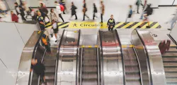 Two STM security officers stand at the top of a bank of escalators as commuters move through a busy metro station concourse, with a yellow 'Circulez' wayfinding stripe visible on the floor above. Two STM security officers stand at the top of a bank of escalators as commuters move through a busy metro station concourse, with a yellow 'Circulez' wayfinding stripe visible on the floor above.