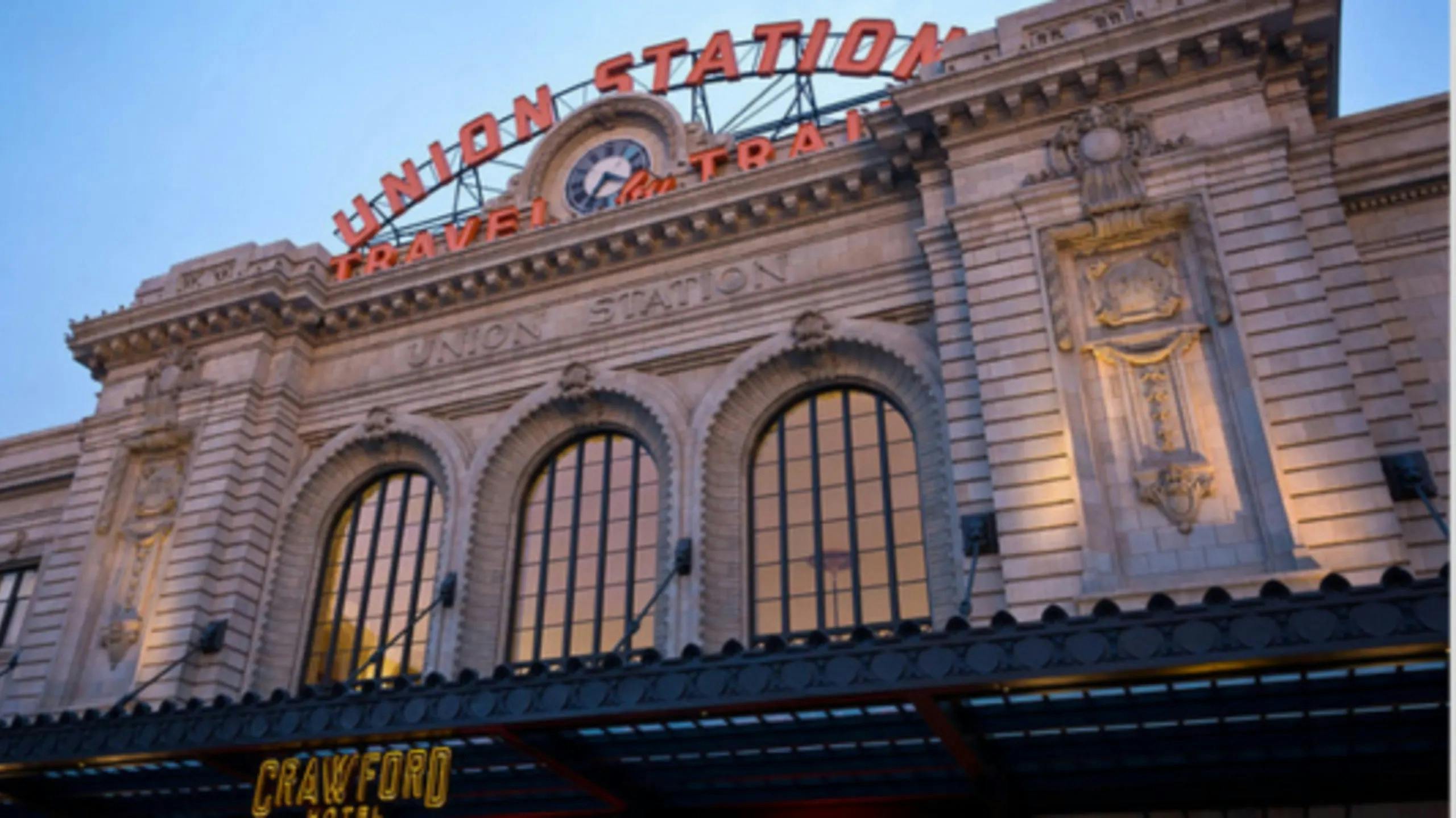The ornate Beaux-Arts facade of Denver Union Station photographed at dusk. The building's cream-colored stone exterior features three large arched windows and decorative stonework. A red neon sign reading 'Union Station / Travel by Train' arches above a clock at the roofline. The Crawford Hotel marquee is visible at the bottom of the frame.