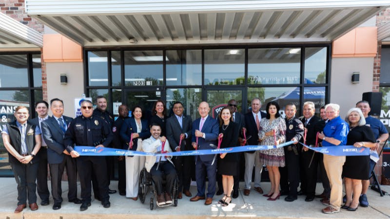 A large group of officials and community members pose for a ribbon-cutting ceremony outside a Metropolitan Transit Authority of Harris County (METRO) facility at 7003 Harrisburg Blvd. Participants hold a blue ribbon printed with the METRO logo. A person using a wheelchair is centered in the front row among the group, which includes uniformed police officers and suited officials.