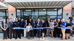 A large group of officials and community members pose for a ribbon-cutting ceremony outside a Metropolitan Transit Authority of Harris County (METRO) facility at 7003 Harrisburg Blvd. Participants hold a blue ribbon printed with the METRO logo. A person using a wheelchair is centered in the front row among the group, which includes uniformed police officers and suited officials.