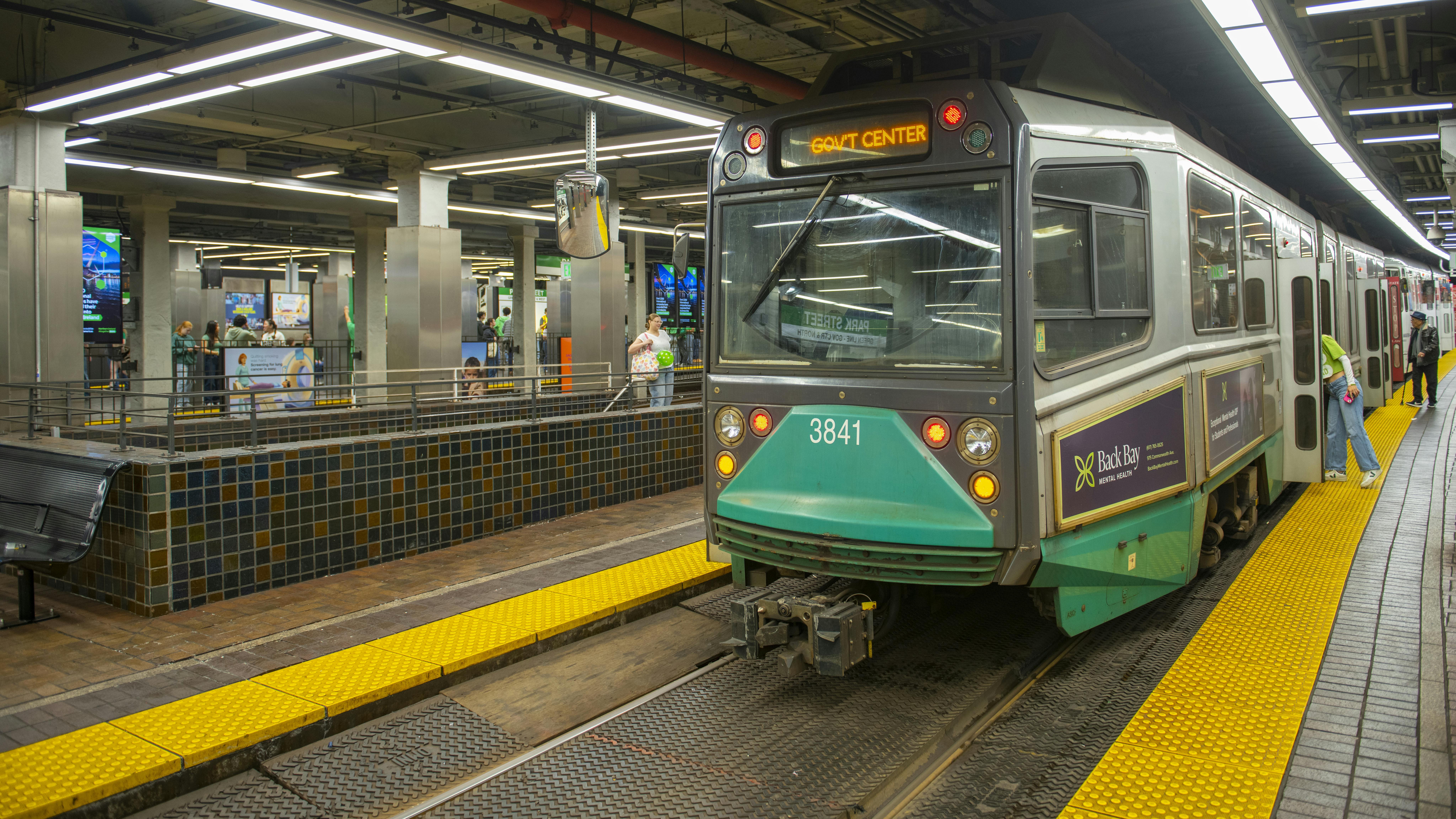 A Massachusetts Bay Transportation Authority Green Line light rail vehicle (car No. 3841), displaying 'Gov't Center' on its destination sign, sits at a station platform. Yellow tactile warning strips line the platform edge in the foreground, while passengers wait and board along the train's length. The underground station features tiled walls, concrete columns, and modern linear LED lighting overhead.