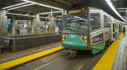 A Massachusetts Bay Transportation Authority Green Line light rail vehicle (car No. 3841), displaying 'Gov't Center' on its destination sign, sits at a station platform. Yellow tactile warning strips line the platform edge in the foreground, while passengers wait and board along the train's length. The underground station features tiled walls, concrete columns, and modern linear LED lighting overhead.