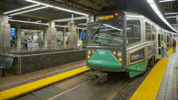 A Massachusetts Bay Transportation Authority Green Line light rail vehicle (car No. 3841), displaying 'Gov't Center' on its destination sign, sits at a station platform. Yellow tactile warning strips line the platform edge in the foreground, while passengers wait and board along the train's length. The underground station features tiled walls, concrete columns, and modern linear LED lighting overhead. A Massachusetts Bay Transportation Authority Green Line light rail vehicle (car No. 3841), displaying 'Gov't Center' on its destination sign, sits at a station platform. Yellow tactile warning strips line the platform edge in the foreground, while passengers wait and board along the train's length. The underground station features tiled walls, concrete columns, and modern linear LED lighting overhead.