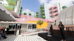 Nicole Ferrara, Deputy Chief Planning Officer, speaks at a podium bearing the Metro 'M' logo during the launch event. Behind her, the new TOD, a multicolored mixed-use residential building stands features green and pink balconies and a bright yellow facade at street level. A sign language interpreter stands to her right, and attendees gather under a tent to the left.