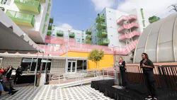 Nicole Ferrara, Deputy Chief Planning Officer, speaks at a podium bearing the Metro 'M' logo during the launch event. Behind her, the new TOD, a multicolored mixed-use residential building stands features green and pink balconies and a bright yellow facade at street level. A sign language interpreter stands to her right, and attendees gather under a tent to the left. Nicole Ferrara, Deputy Chief Planning Officer, speaks at a podium bearing the Metro 'M' logo during the launch event. Behind her, the new TOD, a multicolored mixed-use residential building stands features green and pink balconies and a bright yellow facade at street level. A sign language interpreter stands to her right, and attendees gather under a tent to the left.