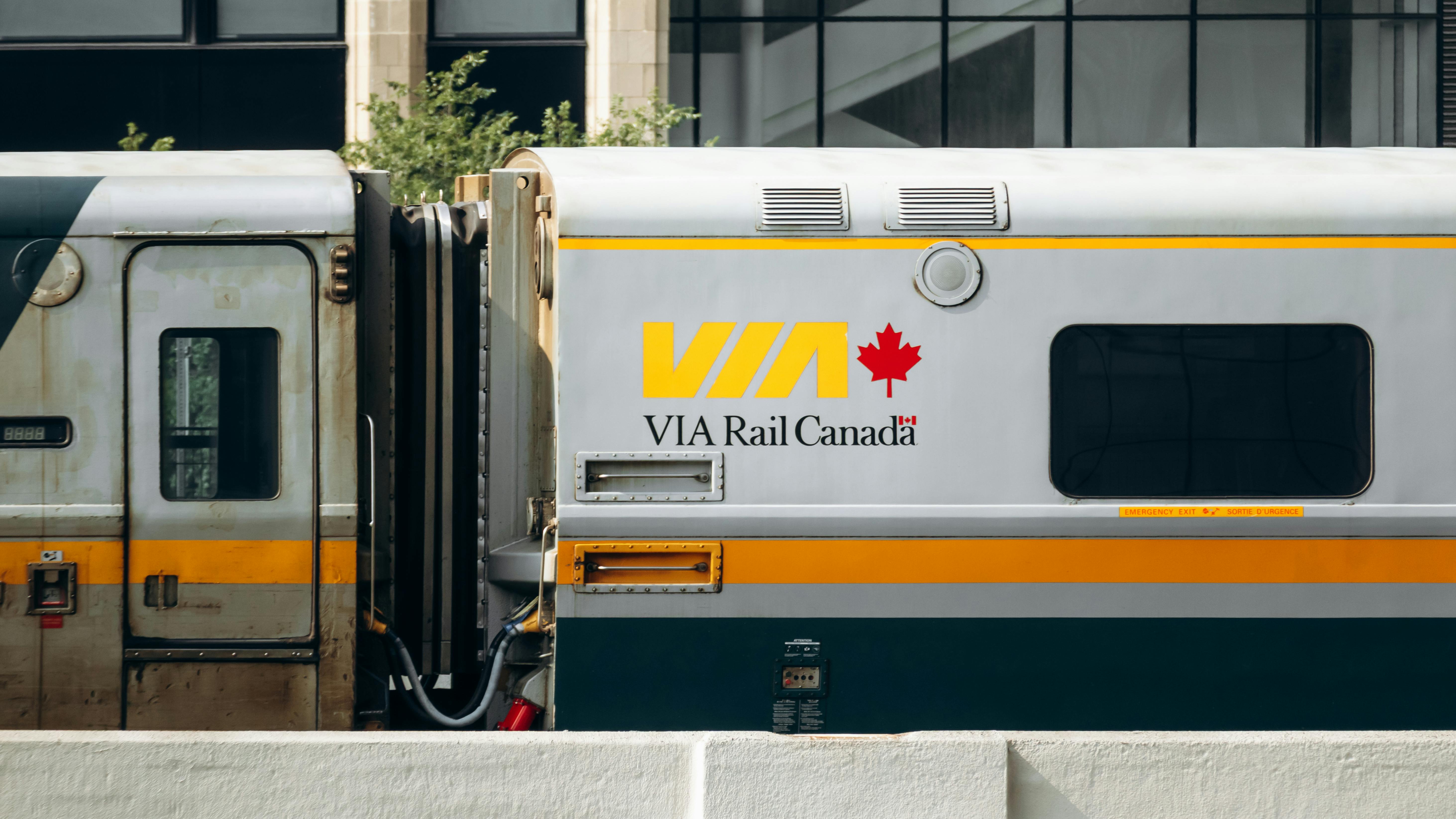 A close-up of a VIA Rail Canada passenger railcar at a station platform. The silver car is accented with yellow and dark teal stripes and displays the VIA logo in bold yellow lettering alongside a red maple leaf. A second, older railcar is visible to the left.
