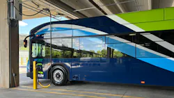 An MCDOT Ride On County Transit electric bus (fleet number 2643E) plugged into a charging pedestal inside a covered depot facility. A yellow charging cable connects the bus to the charger. The bus features a dark blue and light blue wave livery with white accents. A Heliox charging unit is mounted on the wall to the upper left. An MCDOT Ride On County Transit electric bus (fleet number 2643E) plugged into a charging pedestal inside a covered depot facility. A yellow charging cable connects the bus to the charger. The bus features a dark blue and light blue wave livery with white accents. A Heliox charging unit is mounted on the wall to the upper left.