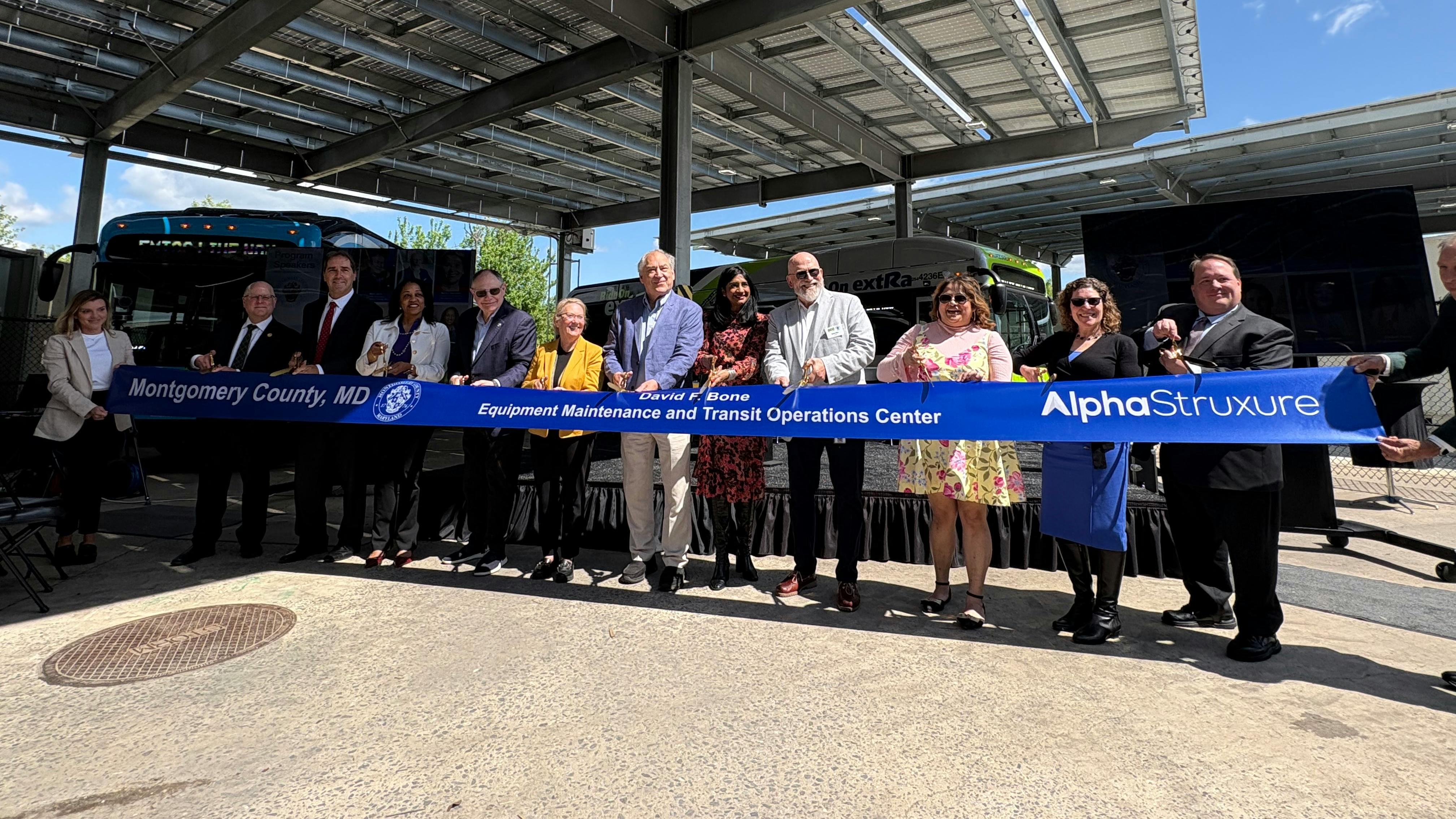 A ribbon-cutting ceremony held under a solar panel canopy at the David F. Bone Equipment Maintenance and Transit Operations Center in Montgomery County, Maryland. Approximately a dozen officials and dignitaries hold a blue ribbon reading 'Montgomery County, MD &mdash; David F. Bone Equipment Maintenance and Transit Operations Center &mdash; AlphaStruxure.' Ride On buses are visible in the background.