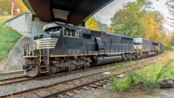 A Norfolk Southern diesel locomotive leads a multi-unit consist under a highway overpass on a double-track mainline corridor. The lead unit is painted in Norfolk Southern's signature black livery with white accent stripes and yellow safety railings. Trees displaying autumn foliage in shades of gold, orange and red line the right-of-way in the background beneath an overcast sky. A Norfolk Southern diesel locomotive leads a multi-unit consist under a highway overpass on a double-track mainline corridor. The lead unit is painted in Norfolk Southern's signature black livery with white accent stripes and yellow safety railings. Trees displaying autumn foliage in shades of gold, orange and red line the right-of-way in the background beneath an overcast sky.
