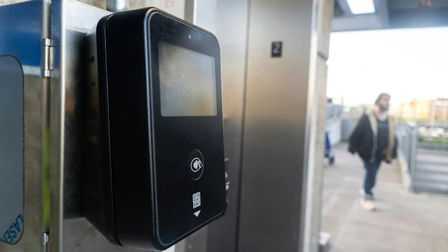 A close-up of a black contactless fare payment terminal mounted on a stainless steel panel at a transit station. The device features a small screen, a tap-to-pay symbol, and a QR code reader. In the background, a blurred figure walks toward a glass-enclosed elevator entrance.