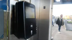 A close-up of a black contactless fare payment terminal mounted on a stainless steel panel at a transit station. The device features a small screen, a tap-to-pay symbol, and a QR code reader. In the background, a blurred figure walks toward a glass-enclosed elevator entrance.