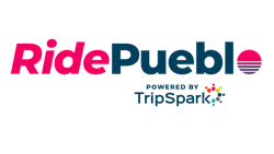 The RidePueblo logo on a transparent background. 'Ride' appears in bold red lettering and 'Pueblo' in dark teal, with a stylized sunset icon replacing the letter 'o' — consisting of a pink semicircle over horizontal red and blue stripes. Below reads 'Powered by TripSpark' with the TripSpark logo, a cluster of colorful dots. The RidePueblo logo on a transparent background. 'Ride' appears in bold red lettering and 'Pueblo' in dark teal, with a stylized sunset icon replacing the letter 'o' — consisting of a pink semicircle over horizontal red and blue stripes. Below reads 'Powered by TripSpark' with the TripSpark logo, a cluster of colorful dots.