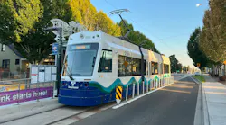 A Sound Transit T Link light rail vehicle, car number 2005, stopped at a street-level station along a tree-lined urban boulevard in Tacoma, Washington. The vehicle is painted white with a blue base and teal wave graphics, with the Sound Transit logo on the nose. A 'Xin Chào' station sign and purple 'Link starts here!' construction banner are visible at left. The photo was taken in warm afternoon light under a clear blue sky.