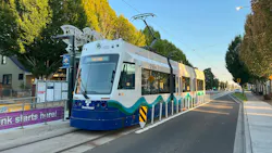 A Sound Transit T Link light rail vehicle, car number 2005, stopped at a street-level station along a tree-lined urban boulevard in Tacoma, Washington. The vehicle is painted white with a blue base and teal wave graphics, with the Sound Transit logo on the nose. A 'Xin Chào' station sign and purple 'Link starts here!' construction banner are visible at left. The photo was taken in warm afternoon light under a clear blue sky. A Sound Transit T Link light rail vehicle, car number 2005, stopped at a street-level station along a tree-lined urban boulevard in Tacoma, Washington. The vehicle is painted white with a blue base and teal wave graphics, with the Sound Transit logo on the nose. A 'Xin Chào' station sign and purple 'Link starts here!' construction banner are visible at left. The photo was taken in warm afternoon light under a clear blue sky.