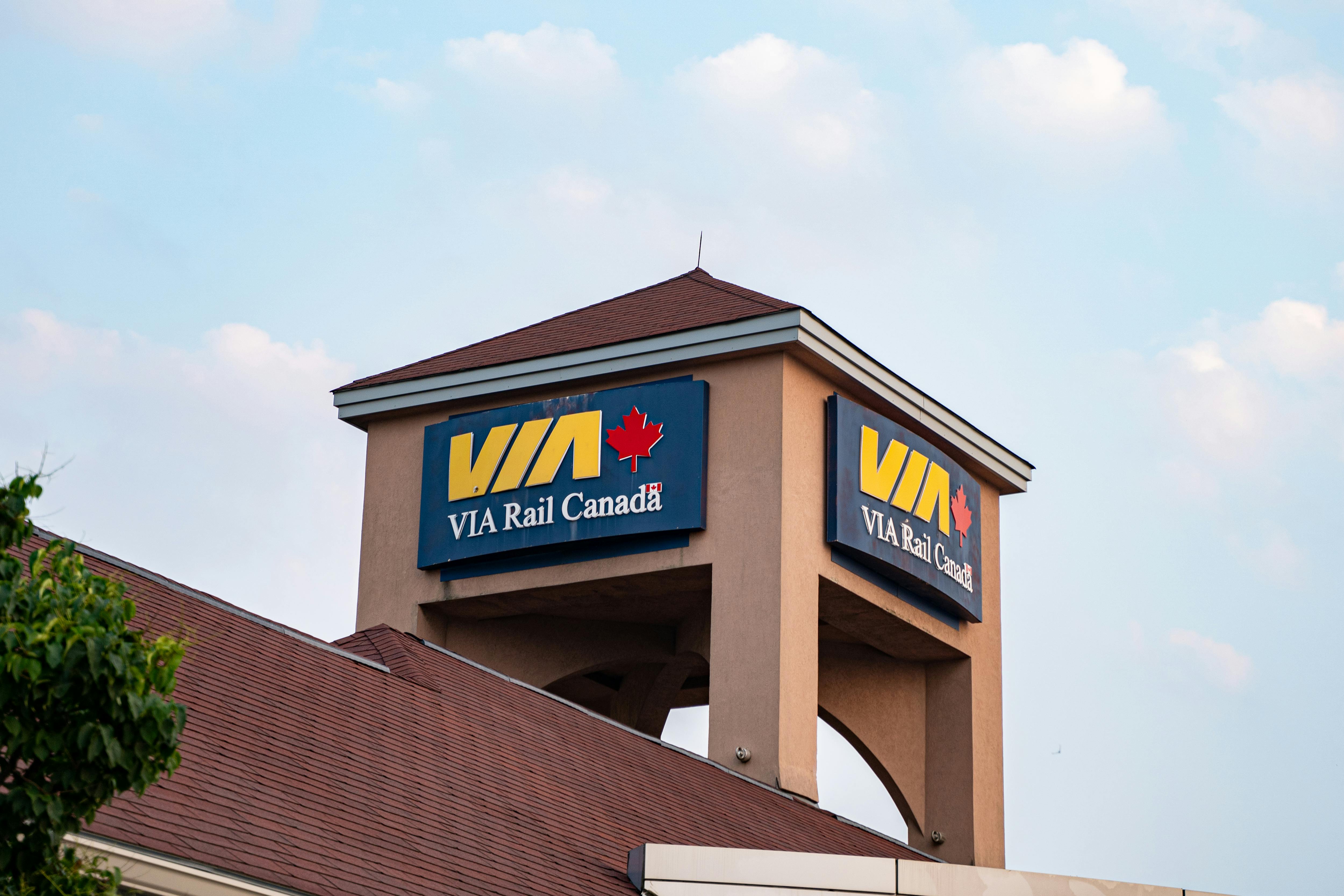 The rooftop tower of a VIA Rail Canada station building, featuring the agency's blue signs with the yellow 'VIA' logo and red maple leaf on two visible sides. The beige stucco tower with a dark red peaked roof rises above the station's sloped roofline against a partly cloudy sky.