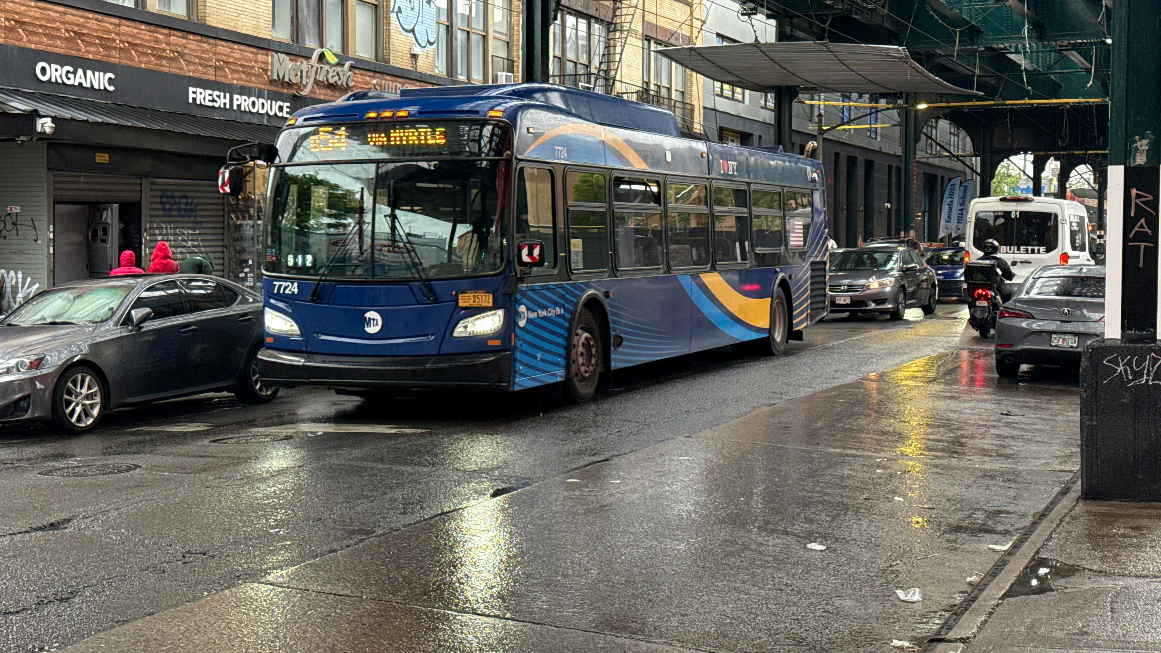 An MTA New York City Bus articulated bus (fleet No. 7724) in navy blue and gold livery travels along a rain-slicked urban street beneath an elevated rail structure, with its destination sign displaying a B54 route toward Myrtle Avenue. Storefronts, graffiti and wet pavement reflect the overcast light in the background.