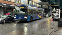 An MTA New York City Bus articulated bus (fleet No. 7724) in navy blue and gold livery travels along a rain-slicked urban street beneath an elevated rail structure, with its destination sign displaying a B54 route toward Myrtle Avenue. Storefronts, graffiti and wet pavement reflect the overcast light in the background. An MTA New York City Bus articulated bus (fleet No. 7724) in navy blue and gold livery travels along a rain-slicked urban street beneath an elevated rail structure, with its destination sign displaying a B54 route toward Myrtle Avenue. Storefronts, graffiti and wet pavement reflect the overcast light in the background.