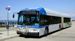 A Community Transit articulated bus (fleet No. 16802) manufactured by New Flyer sits with its destination sign reading 'Out of Service.' The blue, white and gray bus faces forward at a waterfront transit facility on a clear, sunny day, with Puget Sound and distant mountains visible in the background. A bicycle rack is mounted on the front.
