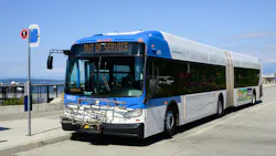 A Community Transit articulated bus (fleet No. 16802) manufactured by New Flyer sits with its destination sign reading 'Out of Service.' The blue, white and gray bus faces forward at a waterfront transit facility on a clear, sunny day, with Puget Sound and distant mountains visible in the background. A bicycle rack is mounted on the front. A Community Transit articulated bus (fleet No. 16802) manufactured by New Flyer sits with its destination sign reading 'Out of Service.' The blue, white and gray bus faces forward at a waterfront transit facility on a clear, sunny day, with Puget Sound and distant mountains visible in the background. A bicycle rack is mounted on the front.