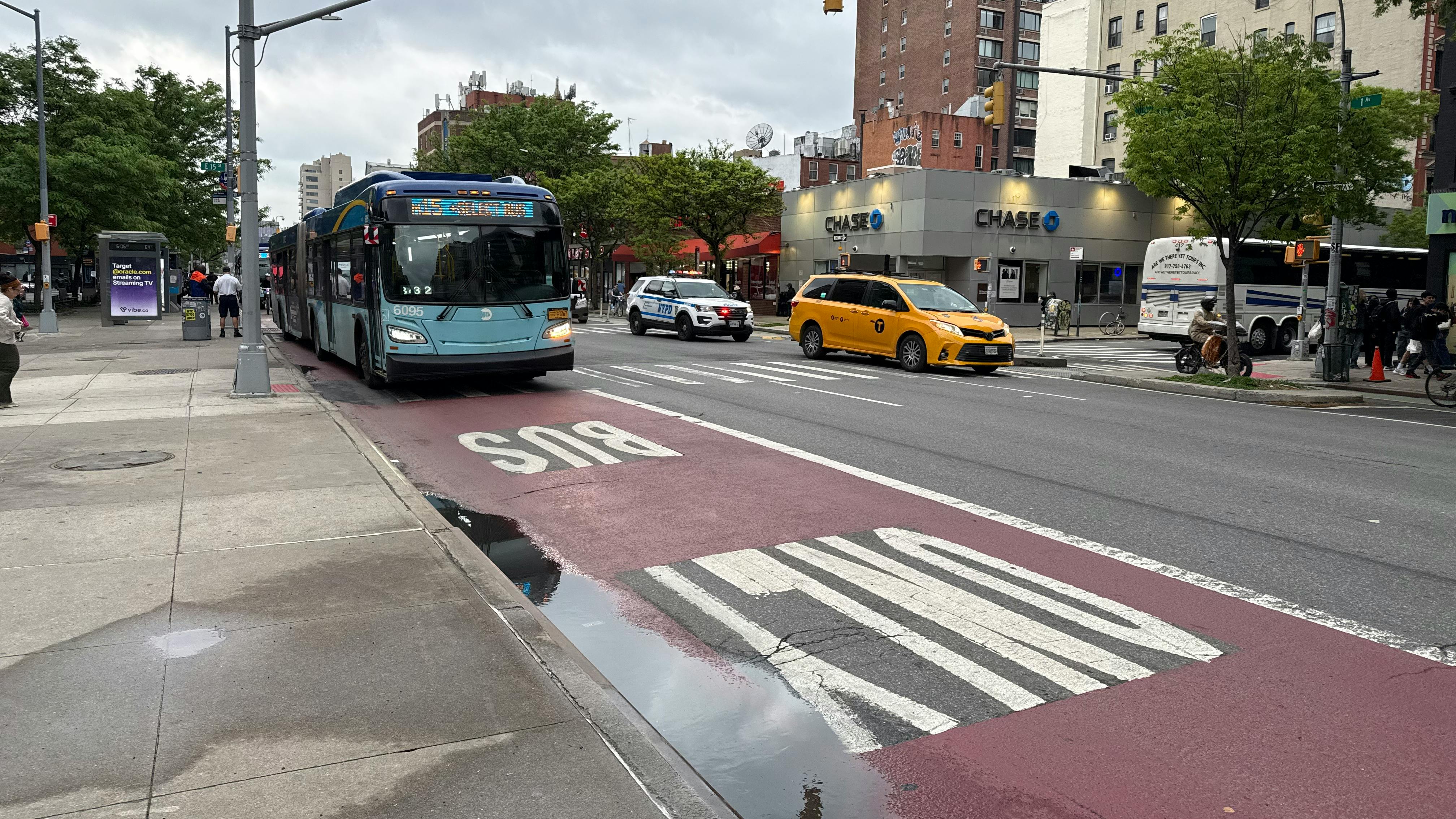 An MTA New York City Bus Select Bus Service articulated bus (fleet No. 6095) travels in a dedicated red bus lane marked 'BUS ONLY' on First Avenue in Manhattan &mdash; a corridor where the MTA has deployed interior-mounted bus lane camera enforcement technology. A yellow taxi, an NYPD vehicle and a Chase bank branch are visible in the background on an overcast day.