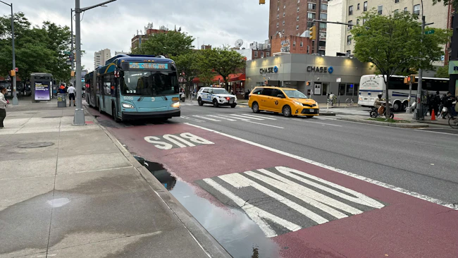 An MTA New York City Bus Select Bus Service articulated bus (fleet No. 6095) travels in a dedicated red bus lane marked 'BUS ONLY' on First Avenue in Manhattan &mdash; a corridor where the MTA has deployed interior-mounted bus lane camera enforcement technology. A yellow taxi, an NYPD vehicle and a Chase bank branch are visible in the background on an overcast day.