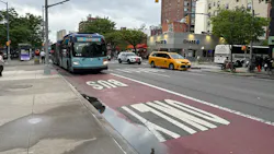 An MTA New York City Bus Select Bus Service articulated bus (fleet No. 6095) travels in a dedicated red bus lane marked 'BUS ONLY' on First Avenue in Manhattan — a corridor where the MTA has deployed interior-mounted bus lane camera enforcement technology. A yellow taxi, an NYPD vehicle and a Chase bank branch are visible in the background on an overcast day. An MTA New York City Bus Select Bus Service articulated bus (fleet No. 6095) travels in a dedicated red bus lane marked 'BUS ONLY' on First Avenue in Manhattan — a corridor where the MTA has deployed interior-mounted bus lane camera enforcement technology. A yellow taxi, an NYPD vehicle and a Chase bank branch are visible in the background on an overcast day.