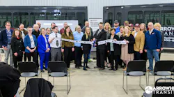 people stand in a line in front of two cutaway buses to cut a ribbon