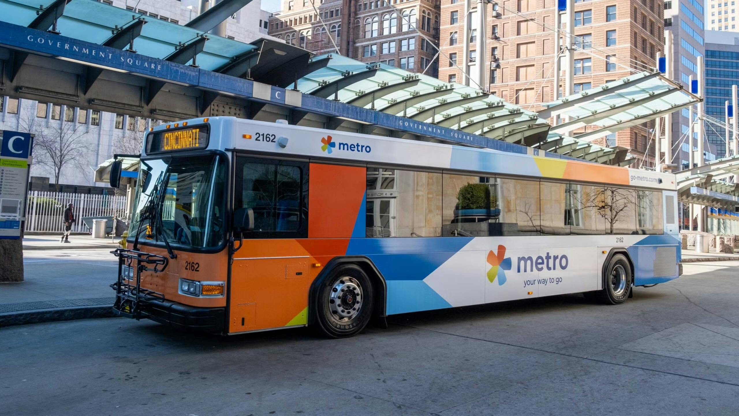 A Metro city bus parked at a downtown transit stop in front of tall office buildings and a glass-covered station platform on a sunny day.