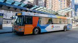 A Metro city bus parked at a downtown transit stop in front of tall office buildings and a glass-covered station platform on a sunny day.