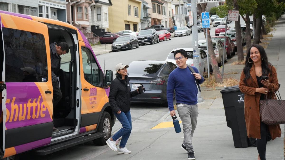 Passengers board and disembark from a purple and orange Bayview Shuttle van on a residential street in San Francisco's Bayview neighborhood. A street sign for Mendell Street is visible in the background.