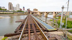 Rail tracks cross a trestle bridge over a river toward a downtown skyline on an overcast day. An arched stone bridge is visible in the background to the right.