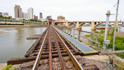 Rail tracks cross a trestle bridge over a river toward a downtown skyline on an overcast day. An arched stone bridge is visible in the background to the right. Rail tracks cross a trestle bridge over a river toward a downtown skyline on an overcast day. An arched stone bridge is visible in the background to the right.
