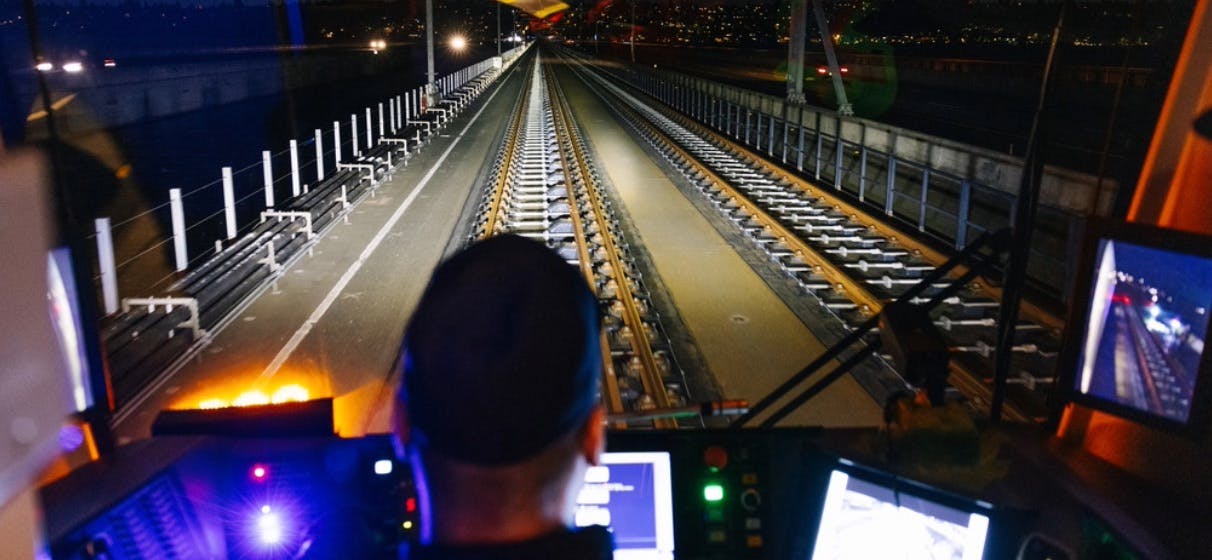 A train operator sits at the controls of a rail vehicle cab, looking ahead down an illuminated elevated track at night. Monitor screens flank the operator on both sides.
