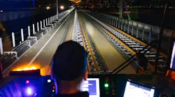 A train operator sits at the controls of a rail vehicle cab, looking ahead down an illuminated elevated track at night. Monitor screens flank the operator on both sides.