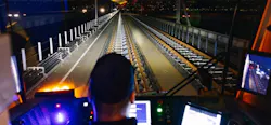 A train operator sits at the controls of a rail vehicle cab, looking ahead down an illuminated elevated track at night. Monitor screens flank the operator on both sides. A train operator sits at the controls of a rail vehicle cab, looking ahead down an illuminated elevated track at night. Monitor screens flank the operator on both sides.