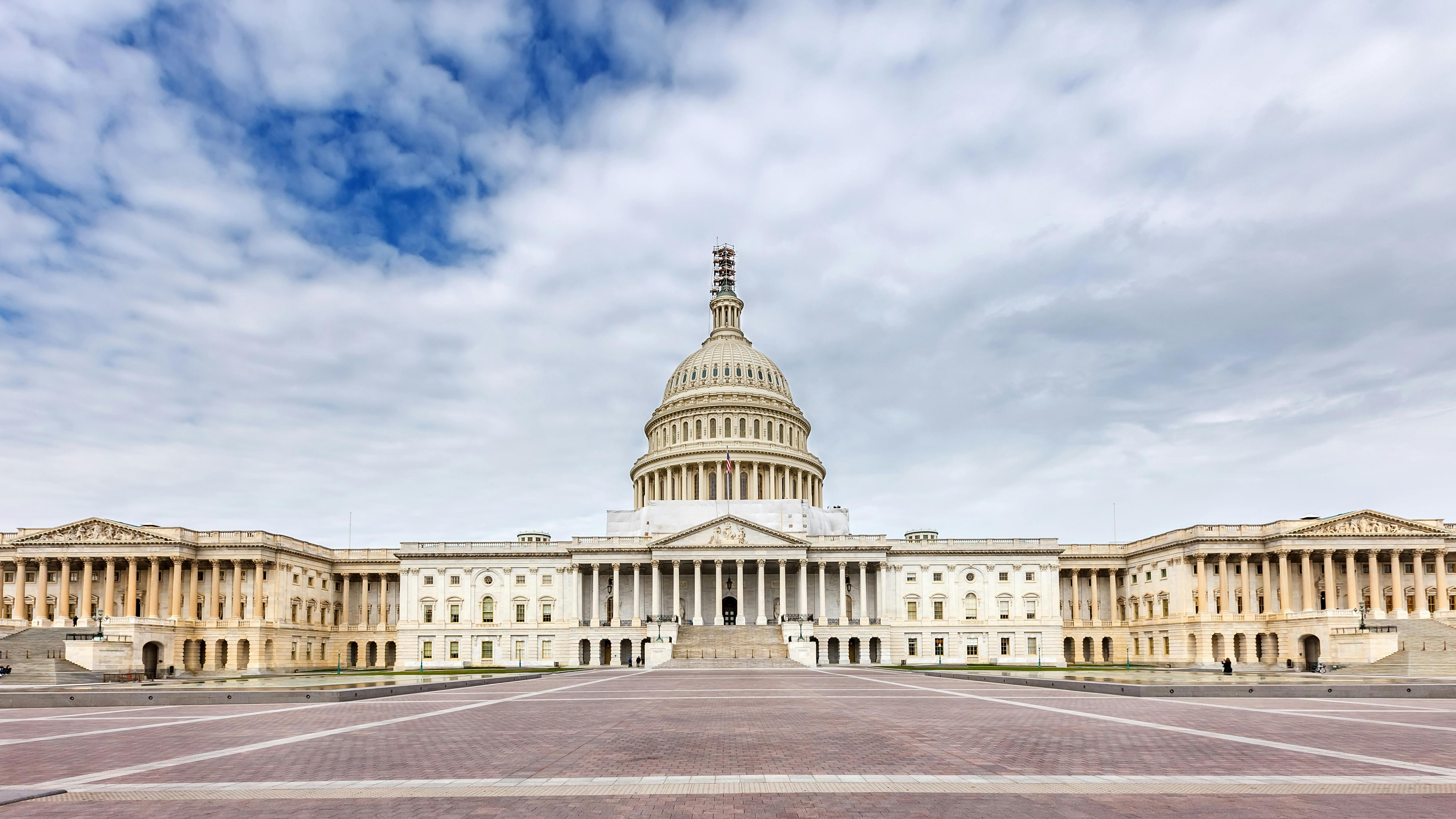 A wide-angle, symmetrical view of the west facade of the U.S. Capitol building in Washington, D.C. The neoclassical structure features a large central dome with scaffolding near the top, flanked by colonnaded wings extending to either side. The foreground shows a broad brick plaza. The sky above is partly cloudy with blue patches visible.