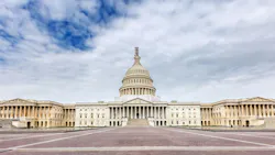 A wide-angle, symmetrical view of the west facade of the U.S. Capitol building in Washington, D.C. The neoclassical structure features a large central dome with scaffolding near the top, flanked by colonnaded wings extending to either side. The foreground shows a broad brick plaza. The sky above is partly cloudy with blue patches visible. A wide-angle, symmetrical view of the west facade of the U.S. Capitol building in Washington, D.C. The neoclassical structure features a large central dome with scaffolding near the top, flanked by colonnaded wings extending to either side. The foreground shows a broad brick plaza. The sky above is partly cloudy with blue patches visible.