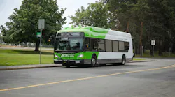 A BC Transit electric bus numbered 7503 travels along a two-lane road with a grassy median and tree line behind it. The bus is white with a bright green front and roof, which features rooftop equipment consistent with a battery-electric or overhead-charging system. The destination sign reads 'Welcome Aboard.' The front carries a bike rack and BC Transit branding.