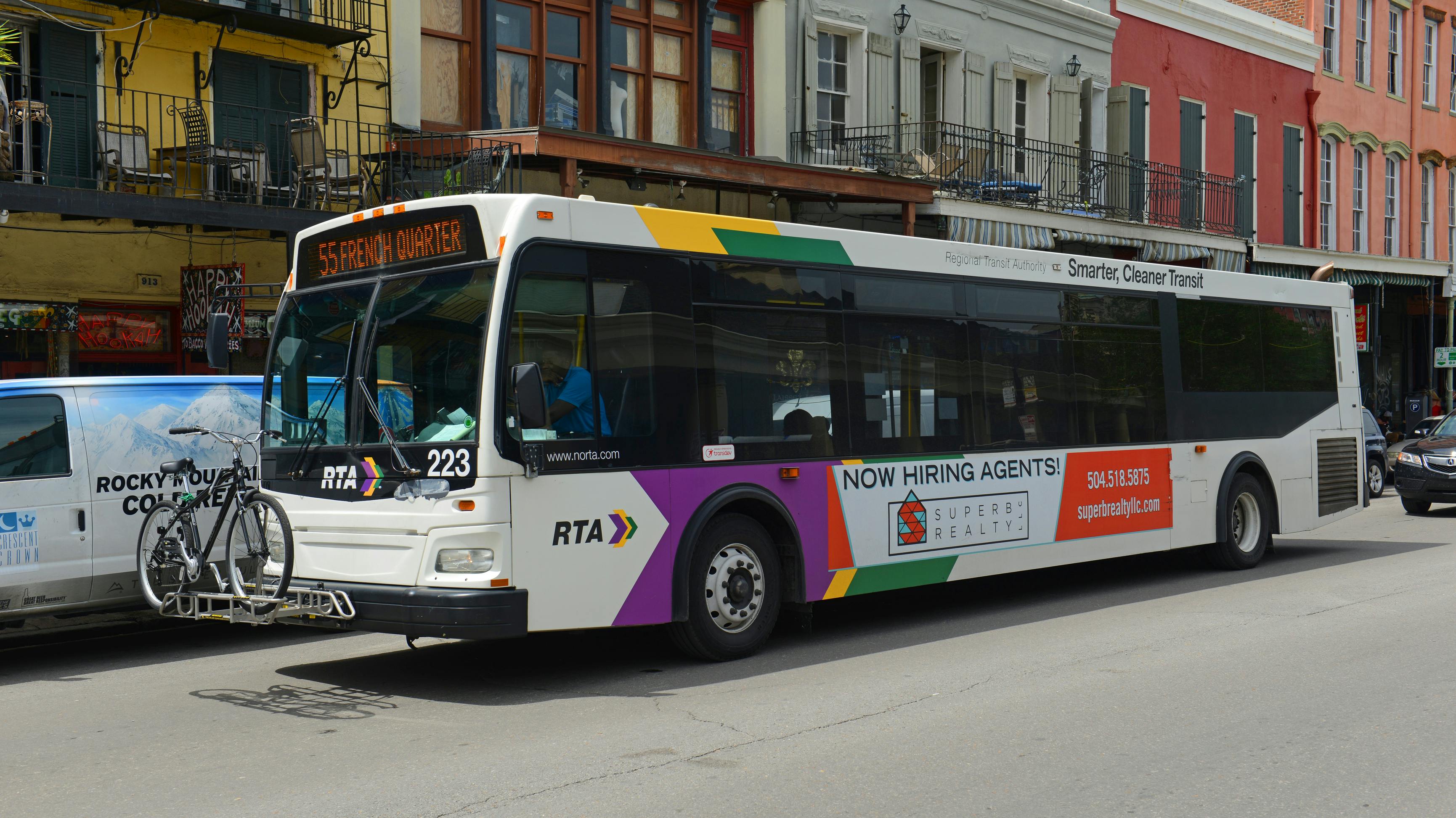 A white New Orleans Regional Transit Authority bus numbered 223 displays '55 French Quarter' on its destination sign as it moves through a street lined with colorful two- and three-story buildings with wrought iron balconies characteristic of New Orleans' French Quarter. The bus features the RTA's multicolor livery of purple, gold, and green accents and carries a bicycle on a front-mounted rack. A side advertisement promotes a realty company. A van is visible to the left.