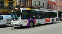 A white New Orleans Regional Transit Authority bus numbered 223 displays '55 French Quarter' on its destination sign as it moves through a street lined with colorful two- and three-story buildings with wrought iron balconies characteristic of New Orleans' French Quarter. The bus features the RTA's multicolor livery of purple, gold, and green accents and carries a bicycle on a front-mounted rack. A side advertisement promotes a realty company. A van is visible to the left.