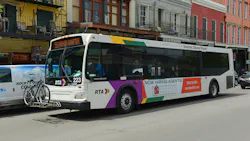 A white New Orleans Regional Transit Authority bus numbered 223 displays '55 French Quarter' on its destination sign as it moves through a street lined with colorful two- and three-story buildings with wrought iron balconies characteristic of New Orleans' French Quarter. The bus features the RTA's multicolor livery of purple, gold, and green accents and carries a bicycle on a front-mounted rack. A side advertisement promotes a realty company. A van is visible to the left. A white New Orleans Regional Transit Authority bus numbered 223 displays '55 French Quarter' on its destination sign as it moves through a street lined with colorful two- and three-story buildings with wrought iron balconies characteristic of New Orleans' French Quarter. The bus features the RTA's multicolor livery of purple, gold, and green accents and carries a bicycle on a front-mounted rack. A side advertisement promotes a realty company. A van is visible to the left.
