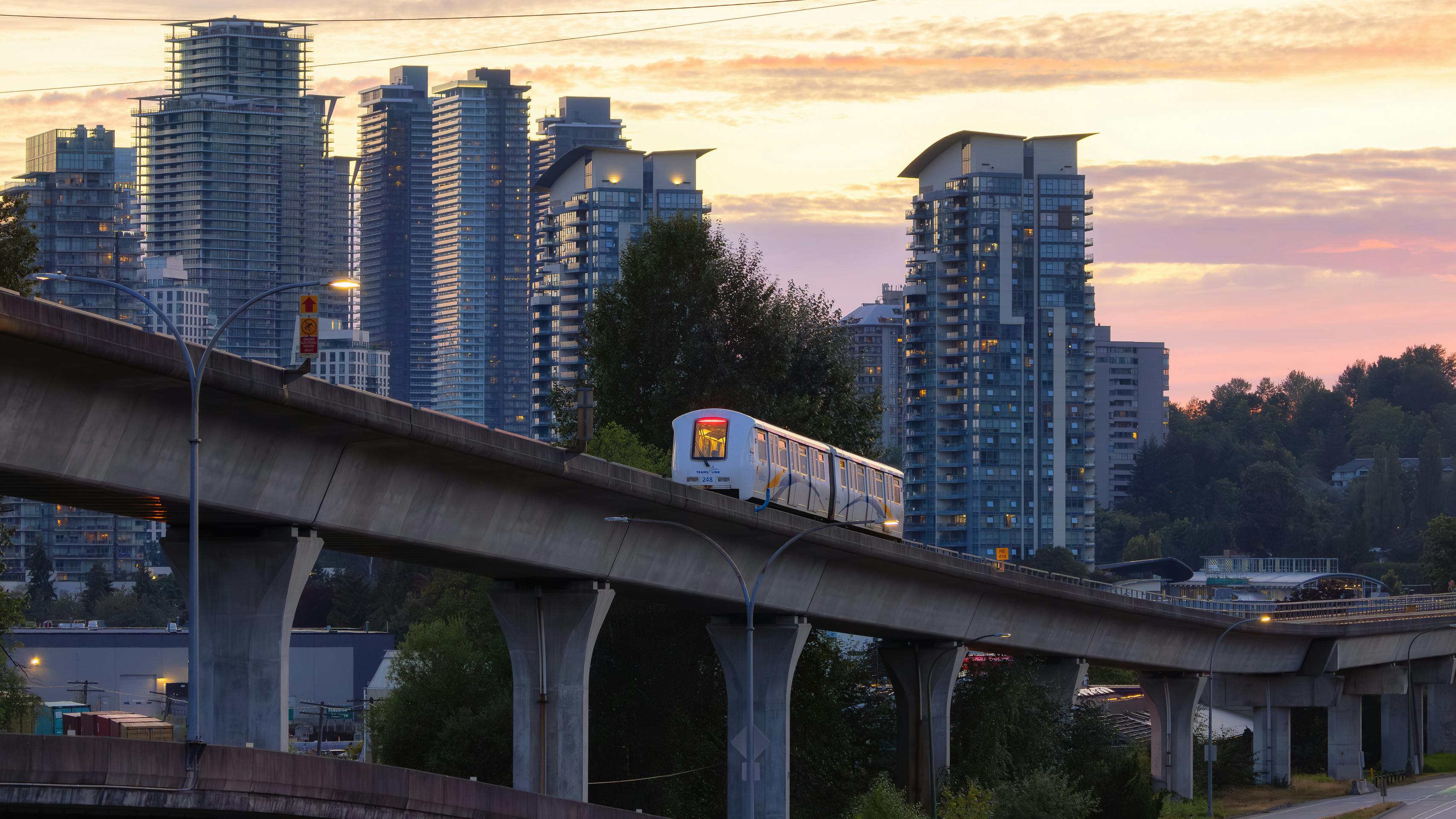 A white TransLink SkyTrain car travels along a curved elevated concrete guideway photographed at dusk. The orange and pink sky silhouettes a dense cluster of modern high-rise residential towers in the background, with mature trees and a roadway visible below. A parallel elevated structure runs along the left side of the frame.