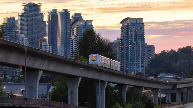 A white TransLink SkyTrain car travels along a curved elevated concrete guideway photographed at dusk. The orange and pink sky silhouettes a dense cluster of modern high-rise residential towers in the background, with mature trees and a roadway visible below. A parallel elevated structure runs along the left side of the frame.