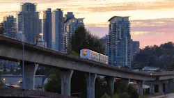 A white TransLink SkyTrain car travels along a curved elevated concrete guideway photographed at dusk. The orange and pink sky silhouettes a dense cluster of modern high-rise residential towers in the background, with mature trees and a roadway visible below. A parallel elevated structure runs along the left side of the frame. A white TransLink SkyTrain car travels along a curved elevated concrete guideway photographed at dusk. The orange and pink sky silhouettes a dense cluster of modern high-rise residential towers in the background, with mature trees and a roadway visible below. A parallel elevated structure runs along the left side of the frame.