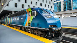 A Tri-Rail locomotive numbered 822 is stopped at a covered station platform with a yellow tactile warning strip along the platform edge. The locomotive wears the railroad's current livery of dark blue with sweeping green, orange and light blue graphic elements. The Tri-Rail wordmark and an RTA logo are visible on the cab. Modern office and mixed-use buildings and a partial view of a palm tree are visible in the background. A Tri-Rail locomotive numbered 822 is stopped at a covered station platform with a yellow tactile warning strip along the platform edge. The locomotive wears the railroad's current livery of dark blue with sweeping green, orange and light blue graphic elements. The Tri-Rail wordmark and an RTA logo are visible on the cab. Modern office and mixed-use buildings and a partial view of a palm tree are visible in the background.